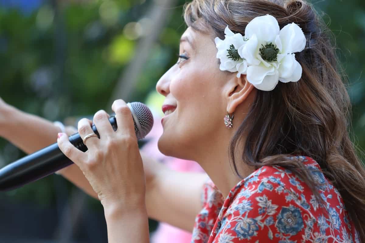 A woman sings into a microphone. She has long brown hair adorned with white flowers tucked behind her ear. She is wearing a red floral patterned top and decorative earrings. The background is out of focus, featuring greenery. Her expression is passionate and engaged.