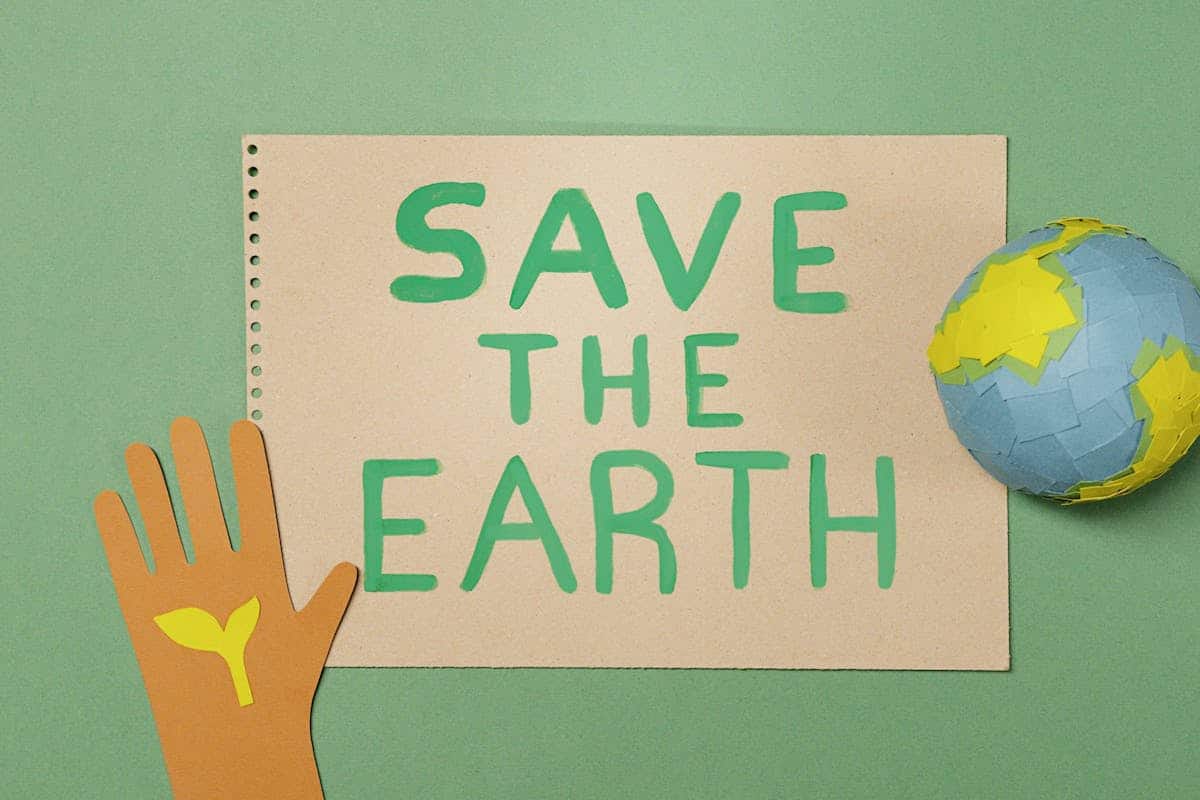 A brown paper cut-out hand with a green sprout symbol sits next to a note reading “SAVE THE EARTH” in green letters, placed on a green background. A small paper globe is beside the note, symbolising environmental conservation. World Earth Day: A Celebration of Our Planet and a Call to Action.